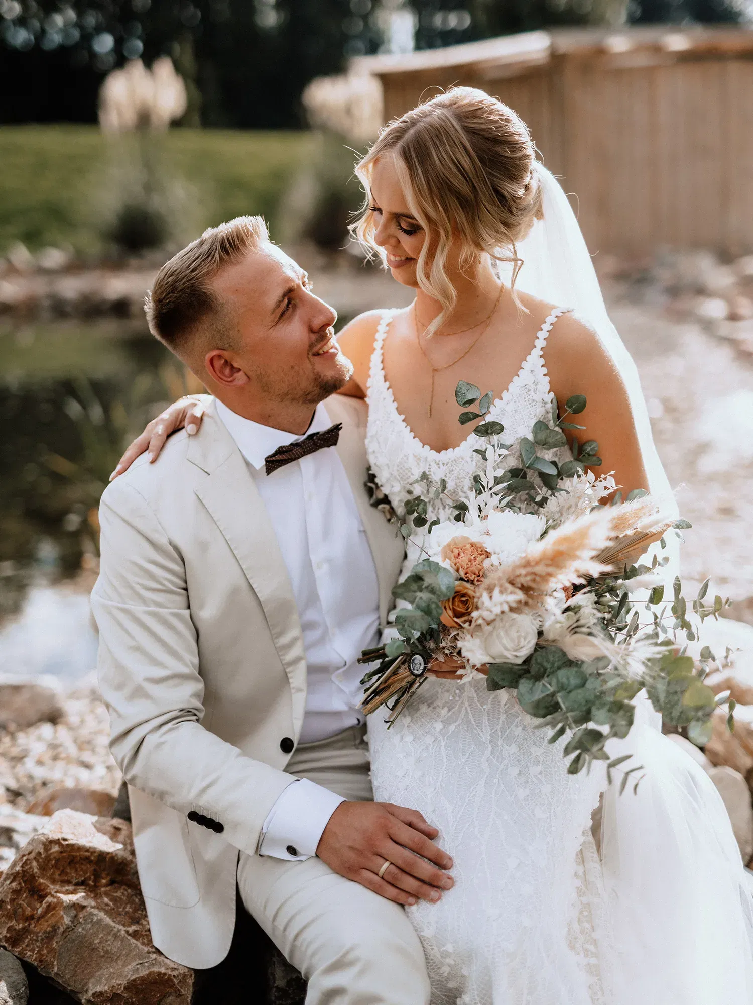 Das Brautpaar genießt einen romantischen Moment im Freien. Die Braut in einem Spitzenkleid und der Bräutigam im cremefarbenen Anzug sitzen am Wasser, umgeben von Natur, während sie sich zärtliche Blicke zuwerfen.