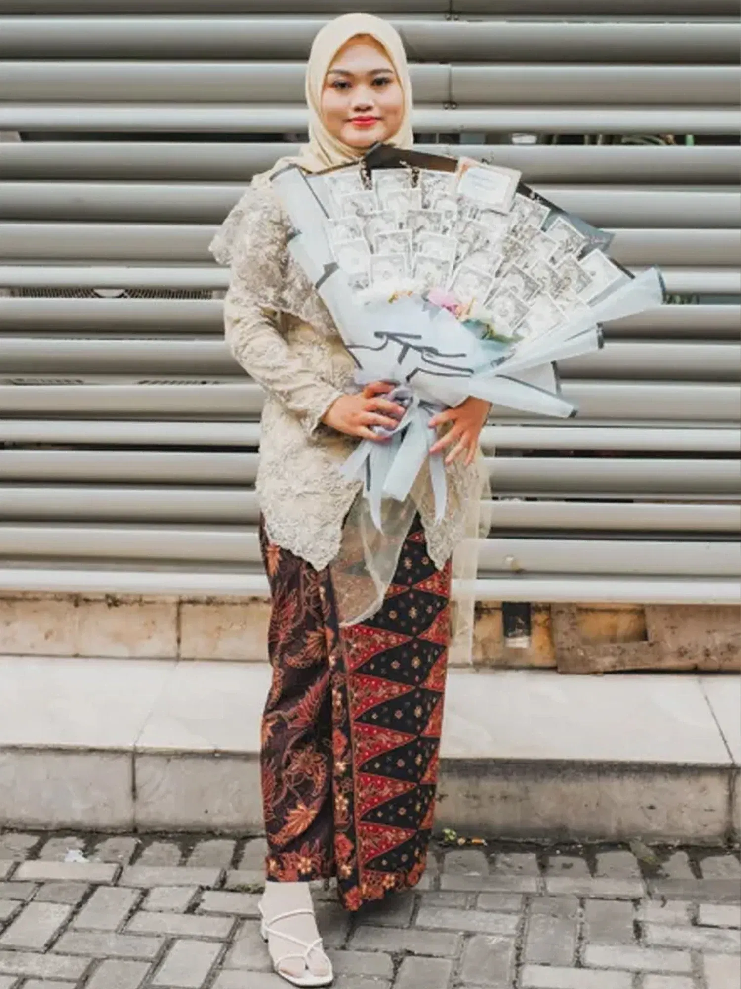 Eine Frau, die ein helles Kopftuch und eine traditionelle Batik-Rock-Kombination trägt, präsentiert einen großen Blumenstrauß, der aus gefalteten Geldscheinen besteht.