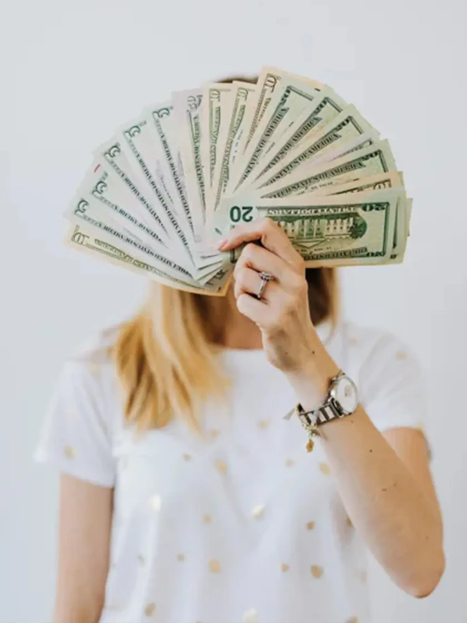 Eine Frau verbirgt ihr Gesicht hinter einem gefächerten Bündel von 20-Dollar-Scheinen. Ihr helles T-Shirt und ihre Uhr sind sichtbar, während die Geldscheine das zentrale Element des Bildes sind.