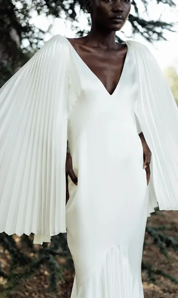 Das Bild zeigt eine stilvolle Braut in einem modernen, elfenbeinfarbenen Kleid mit dramatisch weiten, plissierten Ärmeln, die einen fließenden Effekt erzeugen. Das schlichte Kleid betont den V-Ausschnitt und die klare Linienführung des Designs.