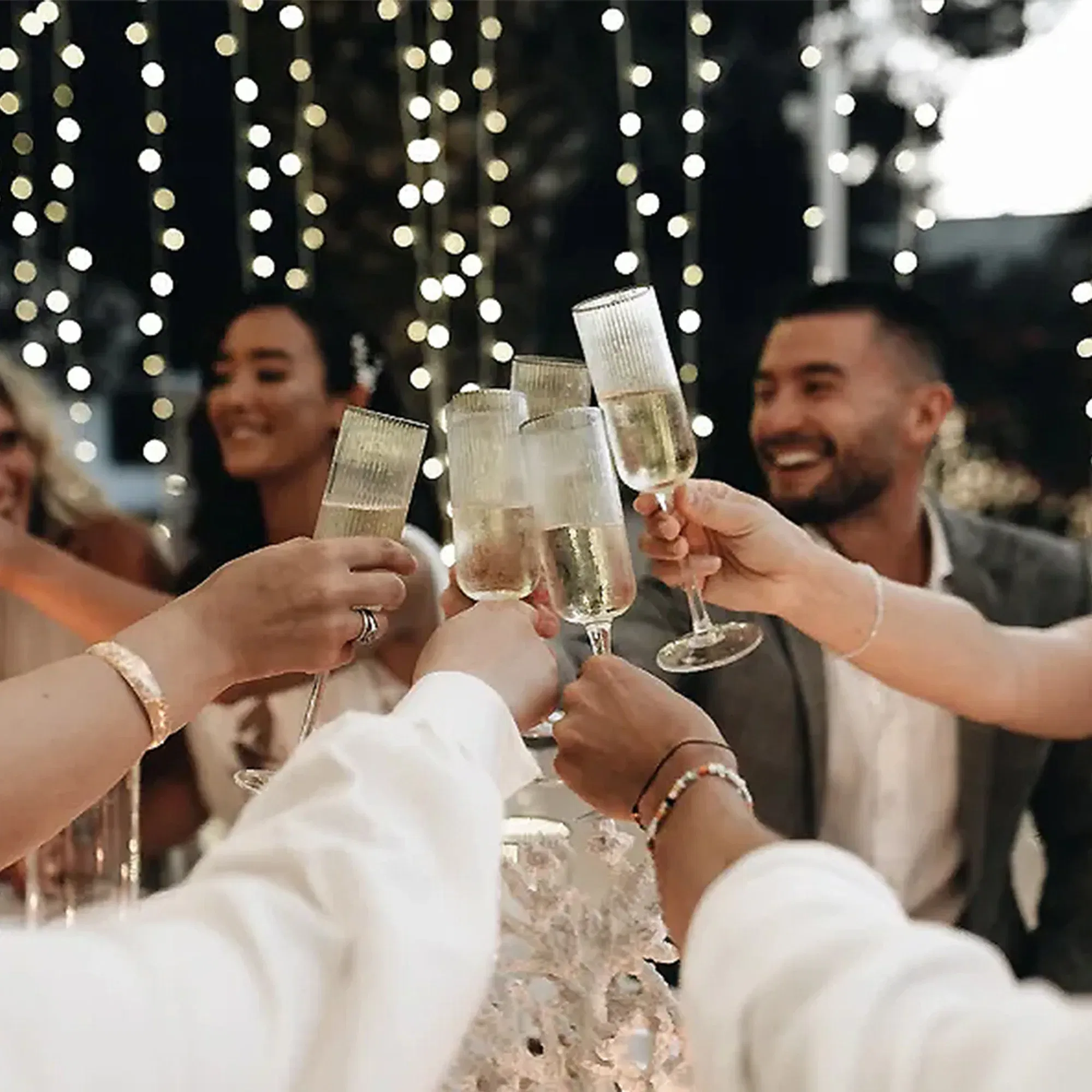 Mehrere Gäste bei einer Hochzeitsfeier heben ihre Sektgläser zu einem gemeinsamen Toast. Der Raum ist von Lichterketten beleuchtet, die eine festliche Stimmung erzeugen. Alle blicken lächelnd und fröhlich in die Runde.