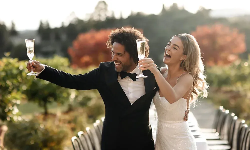 Ein Brautpaar hebt seine Gläser zum Anstoßen inmitten einer grünen Landschaft. Die Braut im weißen Kleid und der Bräutigam im schwarzen Smoking lachen ausgelassen und genießen ihren festlichen Moment.