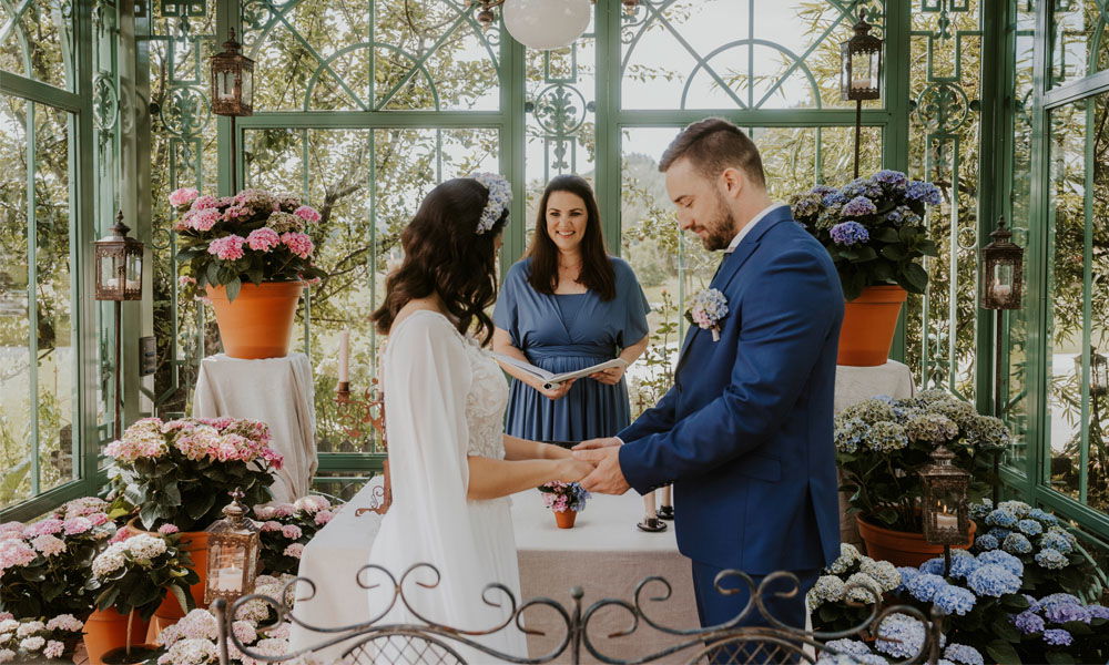 In einem lichtdurchfluteten Gewächshaus mit bunten Hortensien steht ein Brautpaar während ihrer freien Trauung. Die Traurednerin in einem blauen Kleid leitet die Zeremonie.
