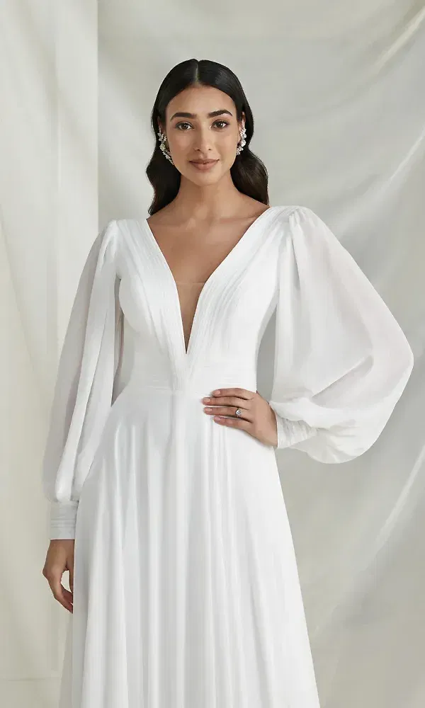 Dieses Bild zeigt eine Braut in einem modernen, minimalistischen Brautkleid mit einem tiefen V-Ausschnitt und langen Puffärmeln. Die klare Linienführung und die sanfte Drapierung des Stoffs verleihen dem Kleid eine raffinierte Eleganz, ideal für eine zeitlose Hochzeitszeremonie.