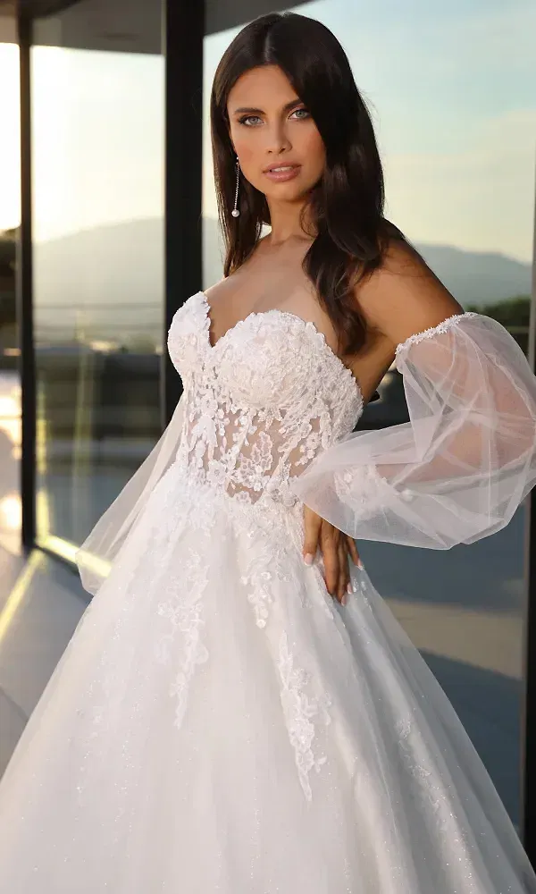 Die Braut trägt ein kunstvoll gearbeitetes, trägerloses Kleid mit transparenten Tüllärmeln und detaillierten Spitzenapplikationen am Oberteil. Sie steht in der Nähe eines Glasgebäudes, im Hintergrund sind sanfte Hügel und der weite Himmel zu sehen, was dem Bild eine elegante, natürliche Atmosphäre verleiht.