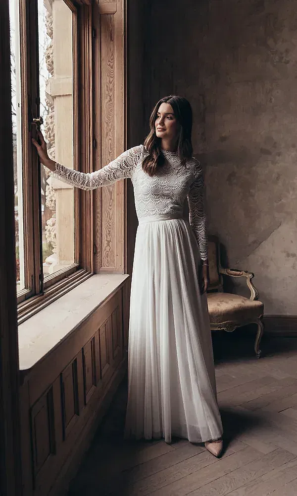 Eine Frau in einem romantischen Brautkleid mit Spitze und einem fließenden Chiffonrock steht nachdenklich am Fenster eines antiken Raumes. Ihr Blick schweift nach draußen, während das warme Tageslicht das filigrane Design ihres Kleides betont.