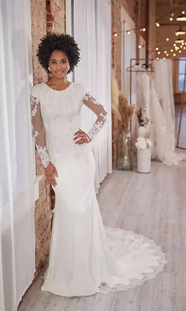 Die Braut trägt ein wunderschönes Spitzenkleid mit langen Ärmeln und steht entspannt in einem modern dekorierten Raum. Das Kleid hat eine elegante Schleppe und betont ihre Figur, während sie in einem charmanten, rustikalen Ambiente posiert.