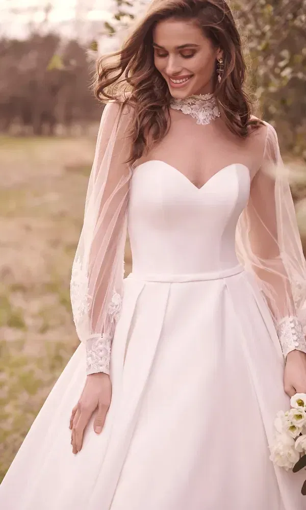 Die Braut trägt ein voluminöses Kleid mit einem Herzausschnitt und durchsichtigen Ärmeln, die mit filigraner Spitze verziert sind. Sie lächelt sanft und steht auf einer Wiese in einem natürlichen Umfeld.