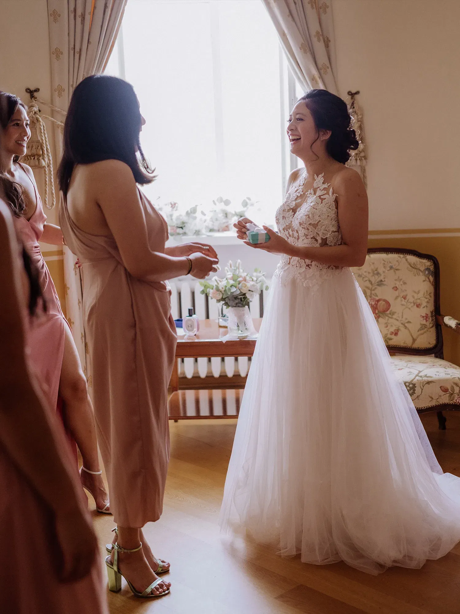 In einem eleganten Zimmer wird die Braut von ihren Freundinnen mit einem Geschenk überrascht. Sie lacht herzlich, während die Brautjungfern, in Pastellkleider gehüllt, sich um sie versammeln.