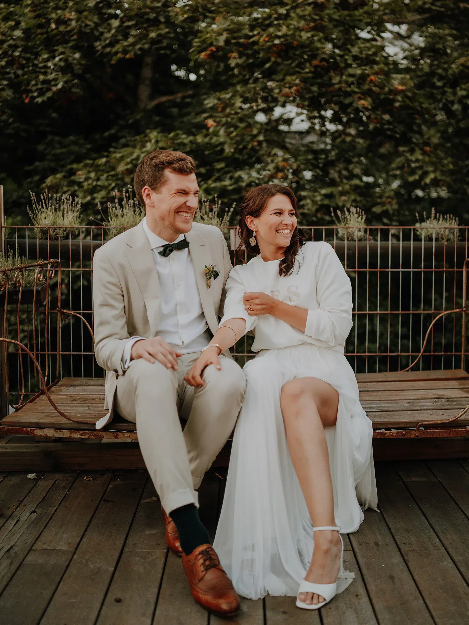 Das frisch verheiratete Paar sitzt fröhlich nebeneinander auf einer Bank im Freien. Die Braut in einem eleganten weißen Kleid und der Bräutigam im beigen Anzug scheinen einen unbeschwerten Moment miteinander zu genießen.