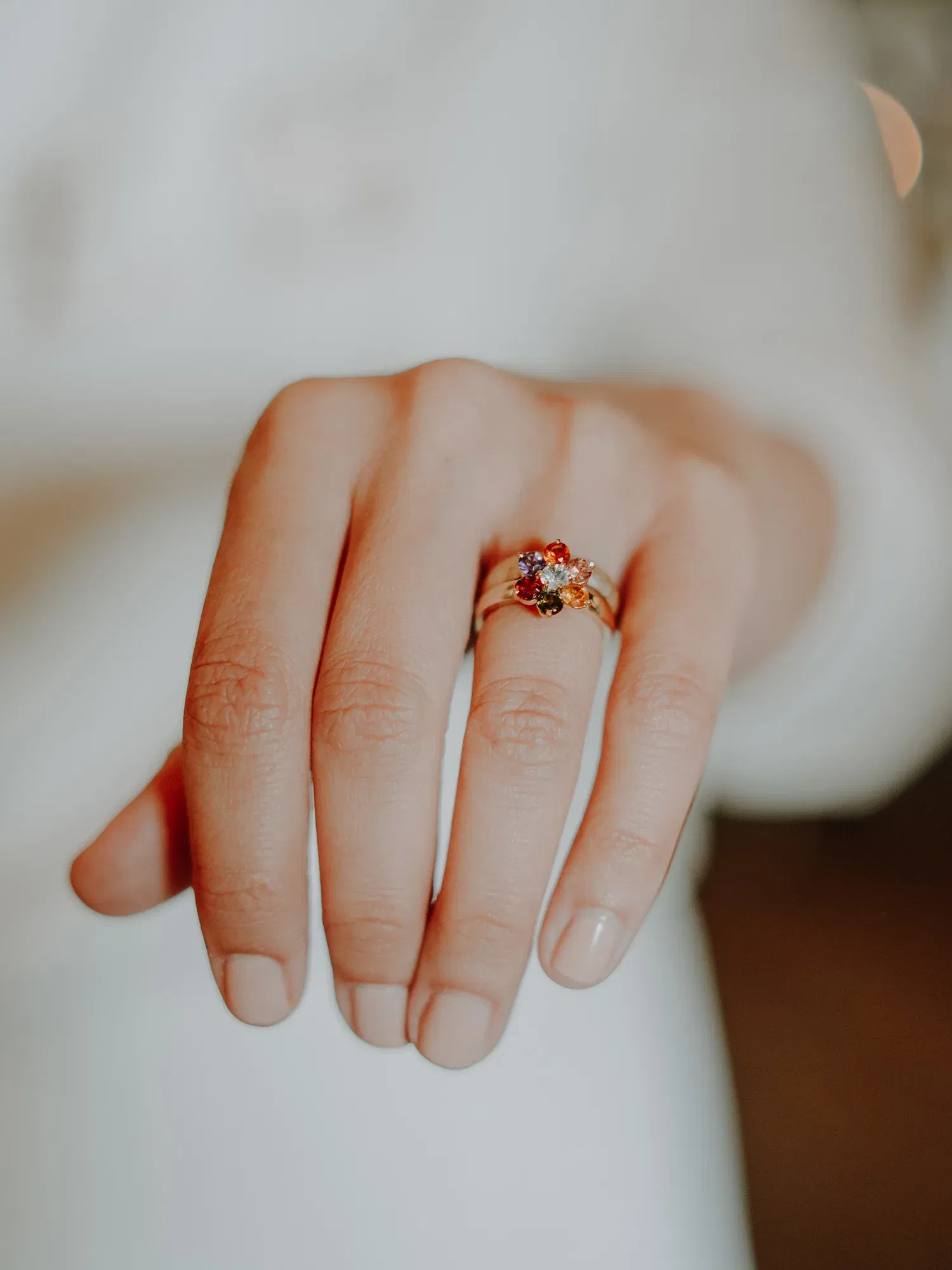 Die Hand einer Person mit dezent gepflegten Nägeln zeigt einen Verlobungsring mit einem leuchtenden Edelstein, der in Gold gefasst ist. Das Bild betont die feinen Details des Rings, während der weiche Hintergrund in Unschärfe gehalten ist.