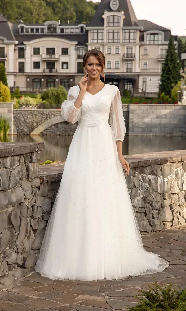 Die Frau trägt ein schlichtes, fließendes Hochzeitskleid mit zarten Ärmeln aus Tüll. Hinter ihr befindet sich ein großes, historisches Gebäude, und sie lehnt sich an eine steinerne Mauer, während sie den Kopf leicht zur Seite neigt.