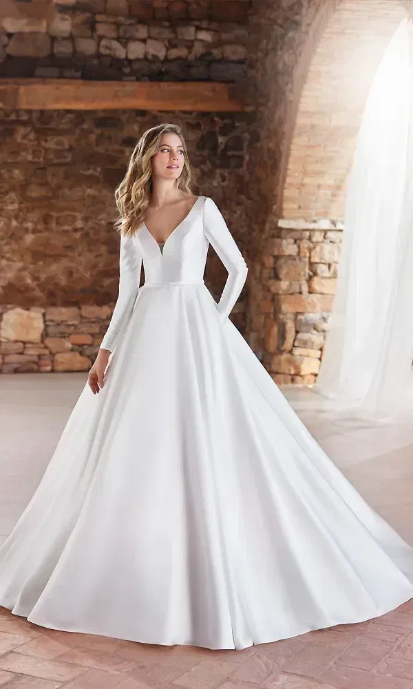 Ein elegantes Hochzeitskleid mit langen Ärmeln, einem tiefen V-Ausschnitt und einem fließenden Rock. Die Braut steht vor einer Steinwand, und das Kleid strahlt schlichte Eleganz und zeitlosen Stil aus, ideal für eine formelle Feier.