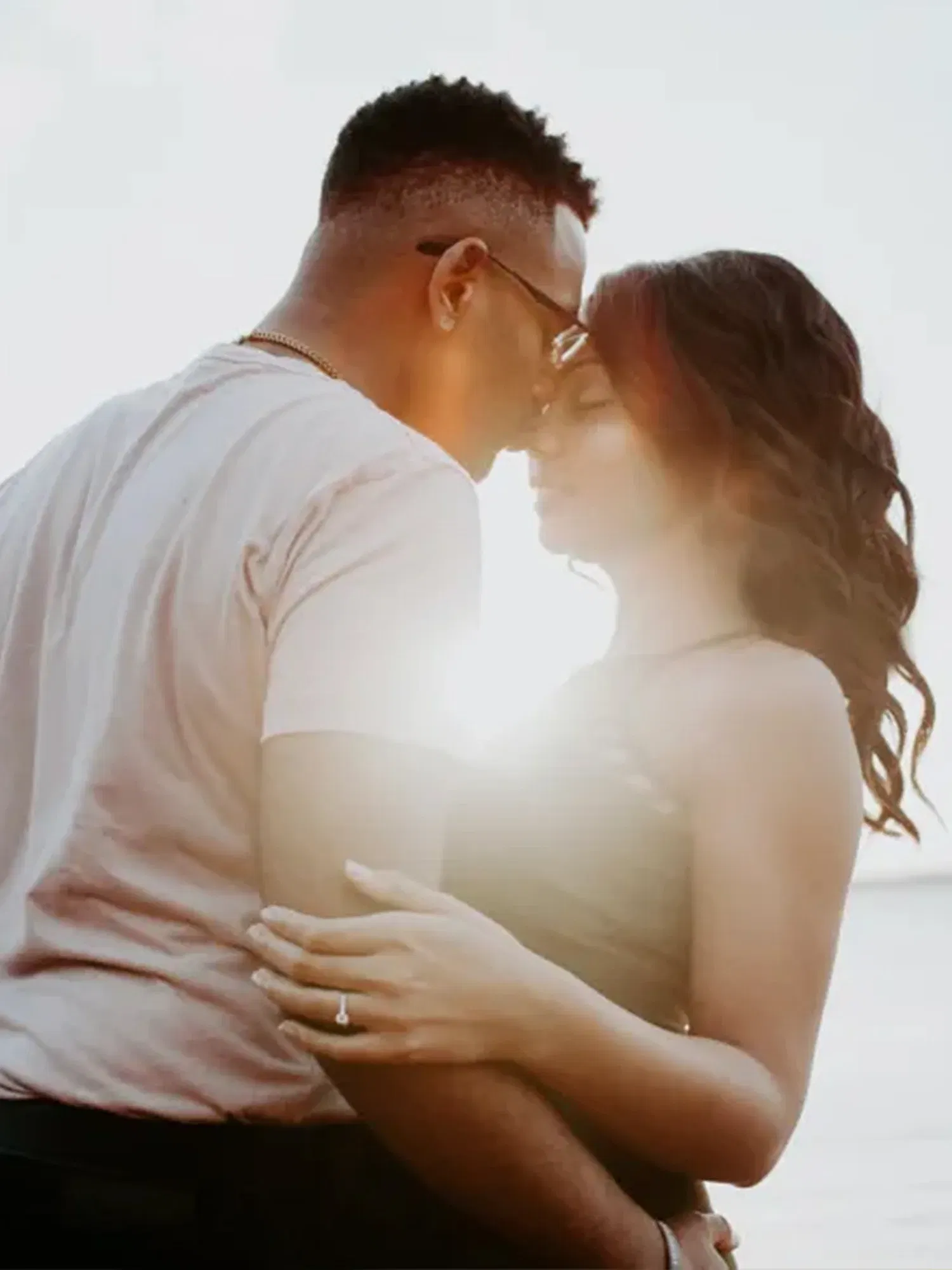 Ein Paar steht sich nahe bei Sonnenuntergang, ihre Stirnen berühren sich sanft. Der Mann hält die Frau zärtlich, während sie die Augen geschlossen hat und ihren Kopf leicht an seinen lehnt. Ihre Hand liegt auf seinem Arm und ein funkelnder Verlobungsring ist gut sichtbar, während die Sonne hinter ihnen leuchtet.