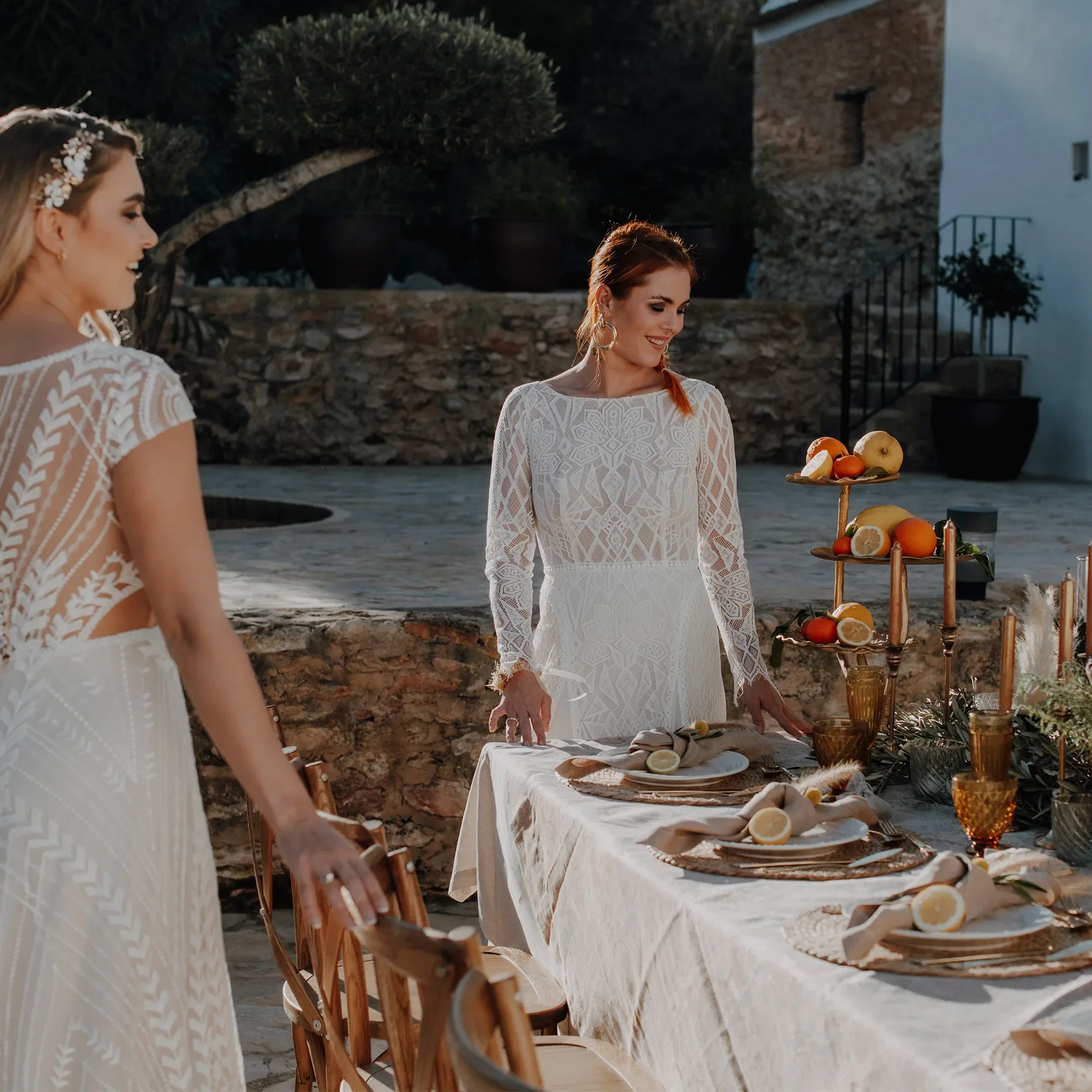 Zwei Bräute in Spitzenkleidern arrangieren eine festliche Tafel mit natürlichen Akzenten und Zitrusfrüchten für eine elegante Outdoor-Hochzeit.