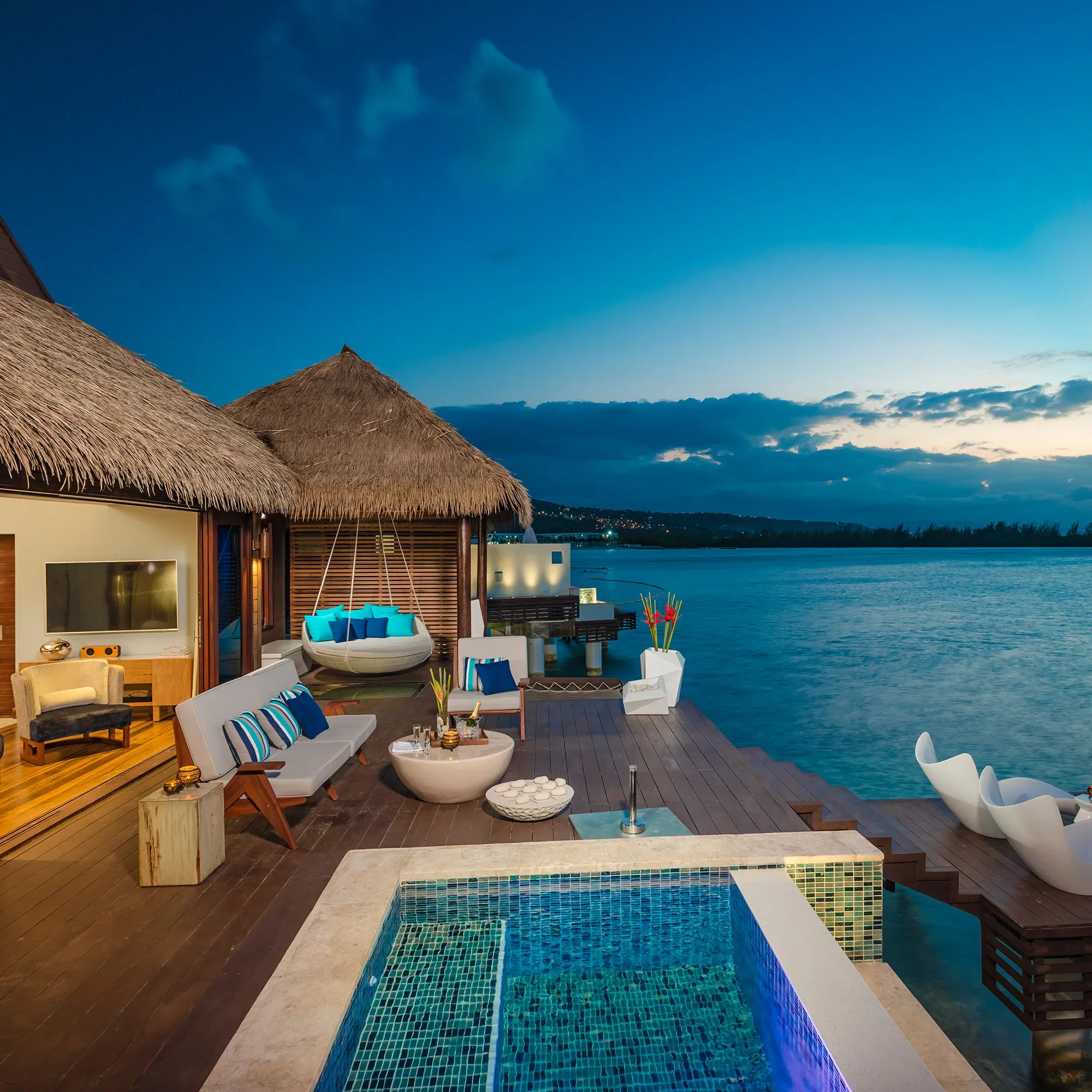 Ein eleganter Überwasser-Bungalow mit Pool und modernen Sitzmöbeln auf einer Holzterrasse, direkt am Wasser gelegen bei Sonnenuntergang.