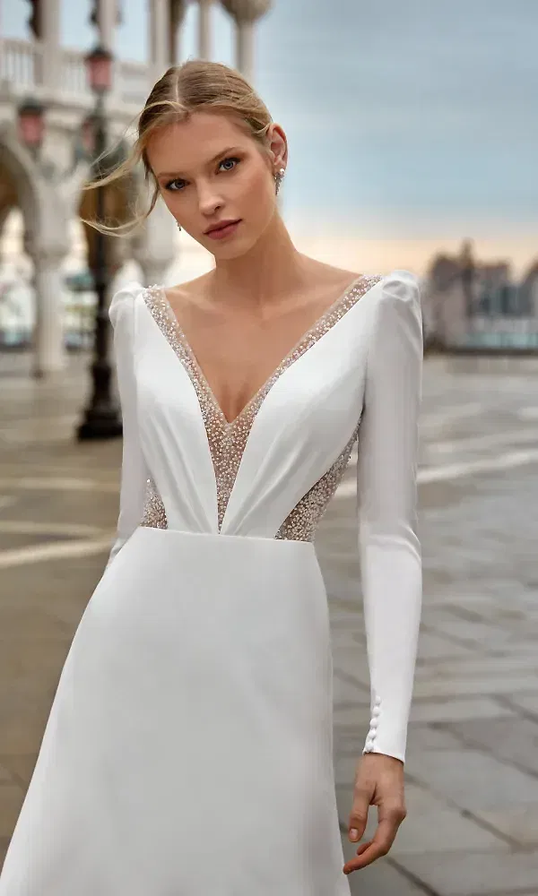 Ein atemberaubendes Hochzeitskleid mit langen Ärmeln und einem tiefen, mit Perlen verzierten V-Ausschnitt. Die Braut steht auf einem stimmungsvollen Platz, das Kleid kombiniert moderne Schlichtheit mit glamourösen Details.