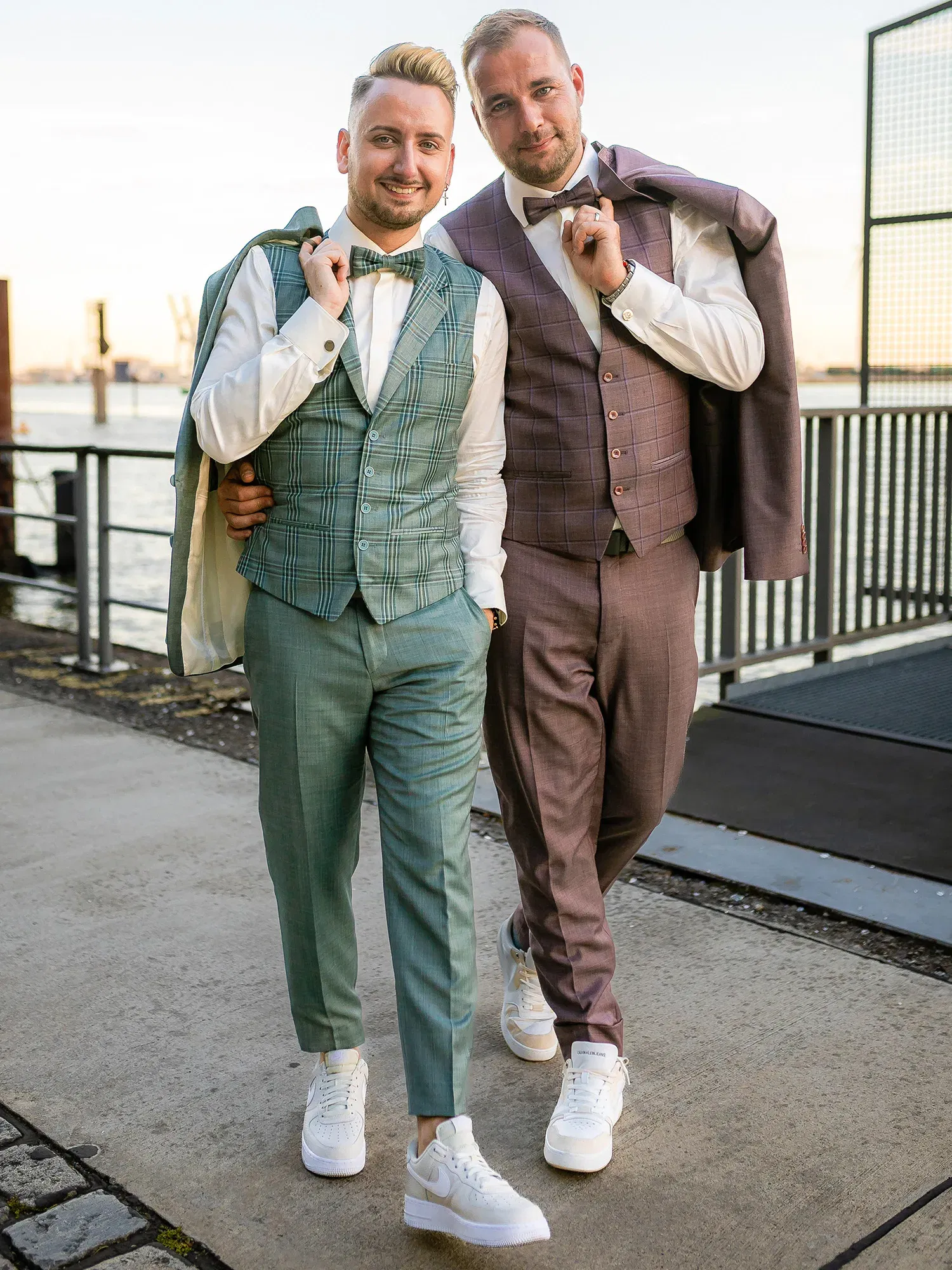 Zwei Männer in modernen, eleganten Anzügen, einer in Grün, der andere in Lila, stehen entspannt nebeneinander an einem Hafengelände. Sie tragen beide weiße Turnschuhe und wirken stilvoll und locker.