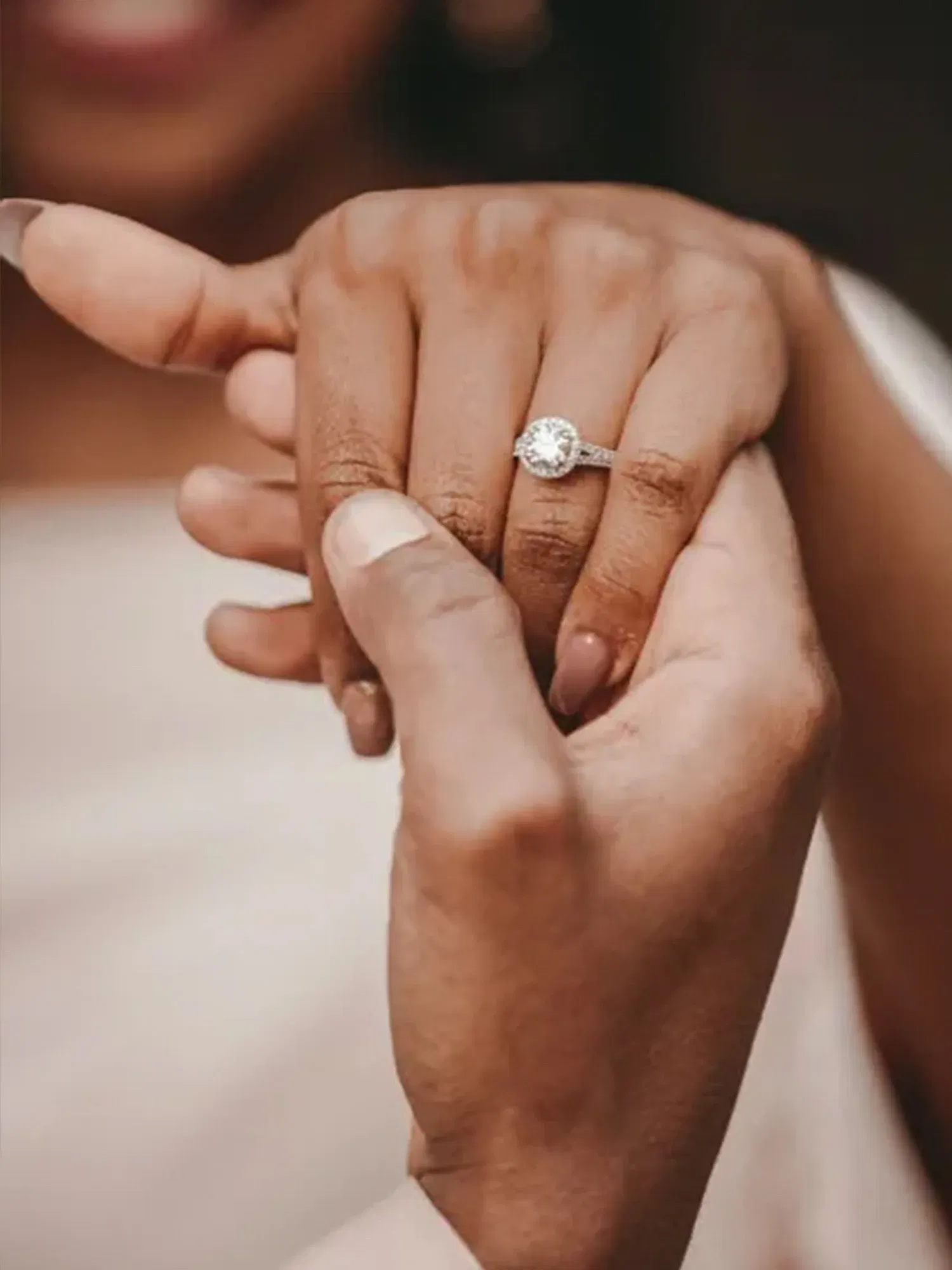 Eine Hand mit einem Verlobungsring, die von einer anderen Hand liebevoll gehalten wird. Der Ring hat einen großen, funkelnden Diamanten, der den Mittelpunkt des Bildes bildet. Der Hintergrund deutet das Lächeln einer Frau an, und die Aufnahme vermittelt Intimität und Freude.