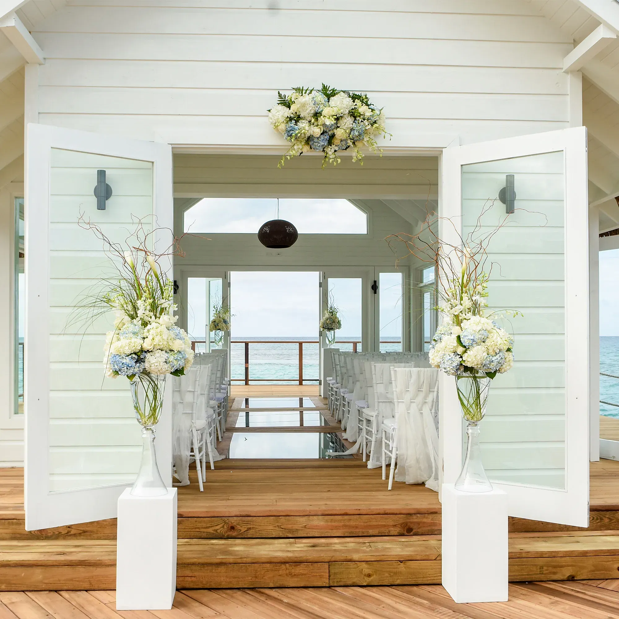 Geöffnete Türen einer weiß gestrichenen Strandkapelle führen zu einem mit Blumen geschmückten Innenraum, dahinter das weite Meer.