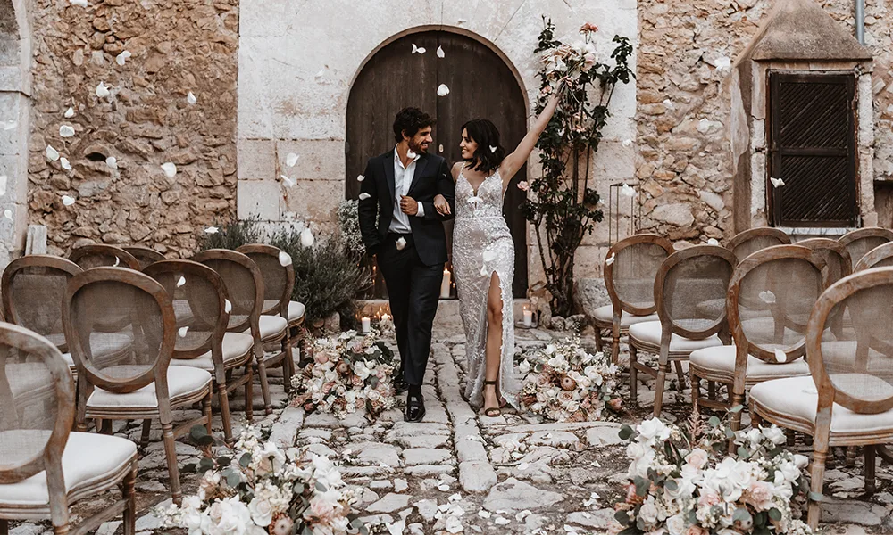 Das frisch verheiratete Paar jubelt und läuft freudestrahlend durch einen mit Blumen und Rosenblättern dekorierten Außenbereich.