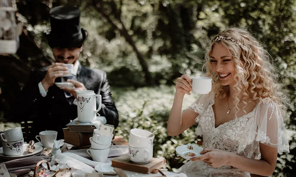 Ein Hochzeitspaar trinkt Tee in einem märchenhaften Setting. Die Braut strahlt vor Freude, während der Bräutigam im Zylinder eine Teetasse hält.