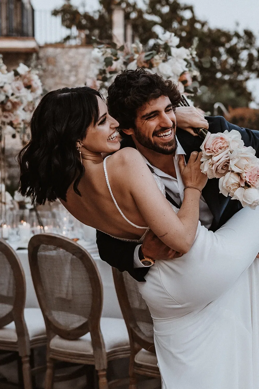 Ein fröhlicher Moment, in dem der Bräutigam seine Braut trägt, beide lachen, während im Hintergrund elegante Hochzeitsdekorationen zu sehen sind.