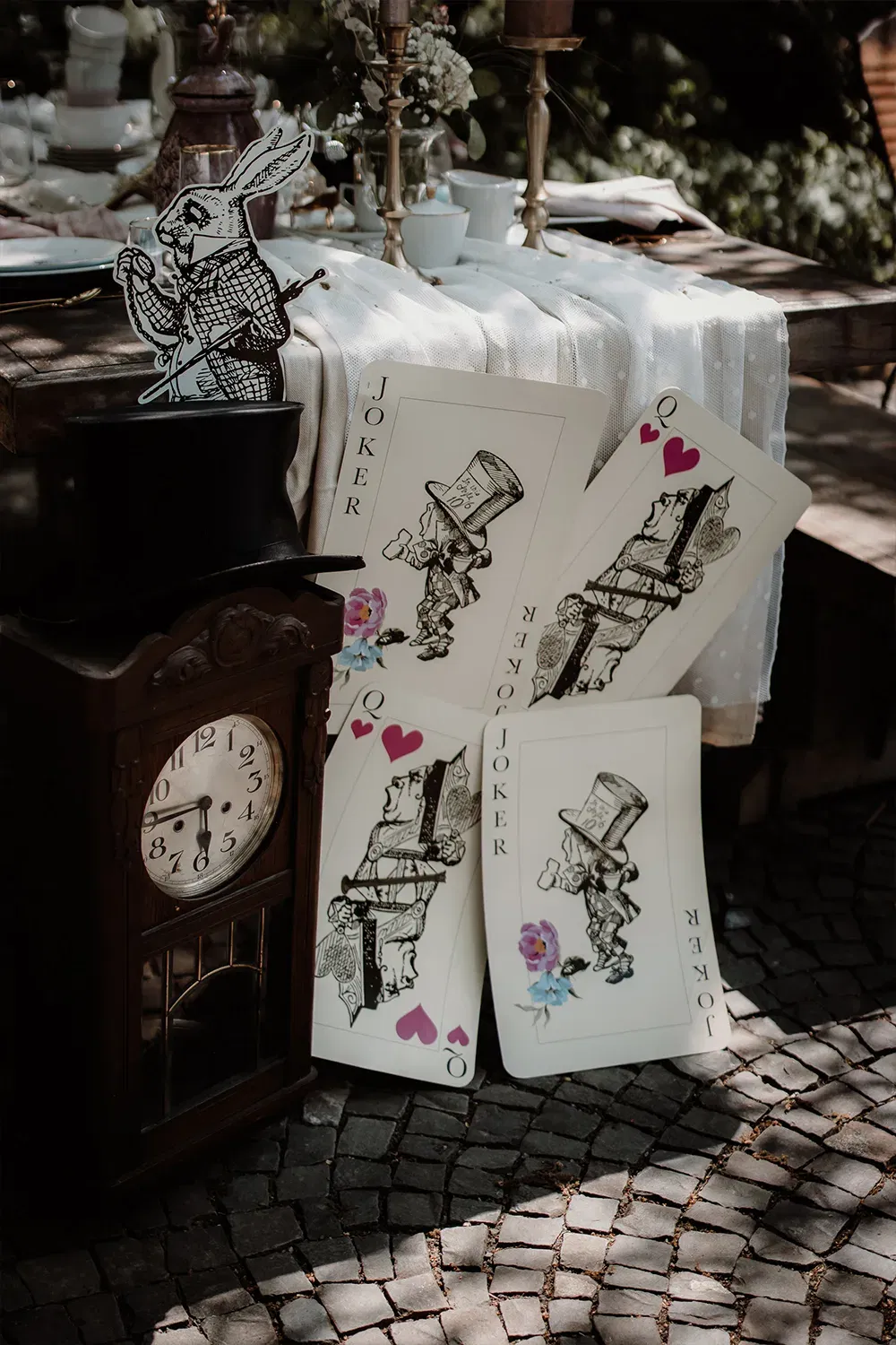 Tischdeko für eine Hochzeit, inspiriert von Alice im Wunderland, mit großen Joker-Karten, einer alten Standuhr und dem weißen Kaninchen als Dekoration.