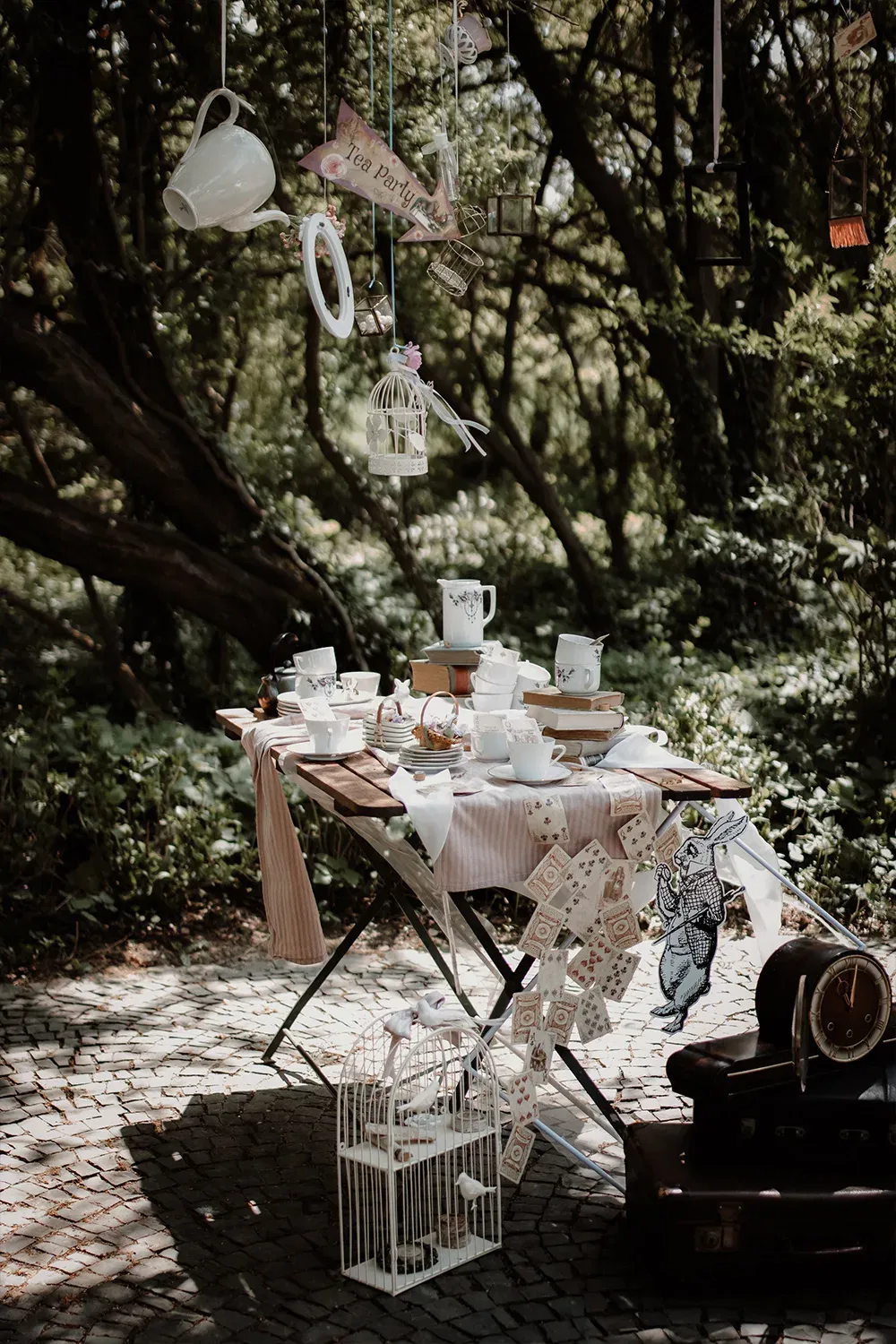 Ein liebevoll dekorierter Outdoor-Teetisch mit gestapeltem Porzellan und fliegenden Teekannen, umgeben von Bäumen in einem Wald.