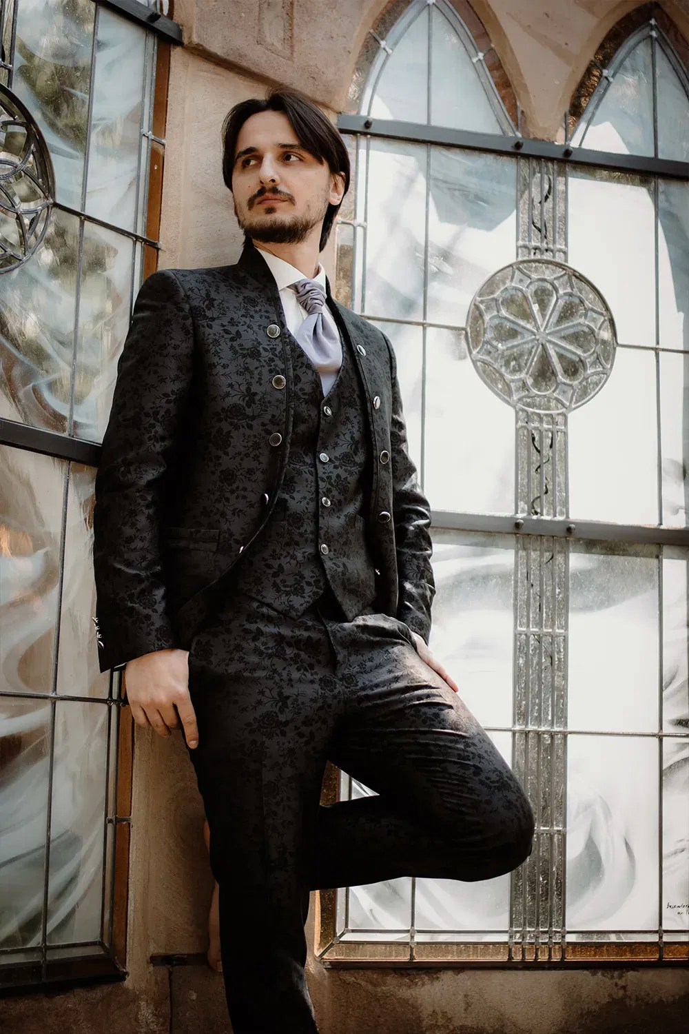 Der Bräutigam, gekleidet in einen schwarzen Anzug mit floralen Mustern, steht entspannt vor einer Glasfenster-Kulisse und schaut nachdenklich zur Seite.