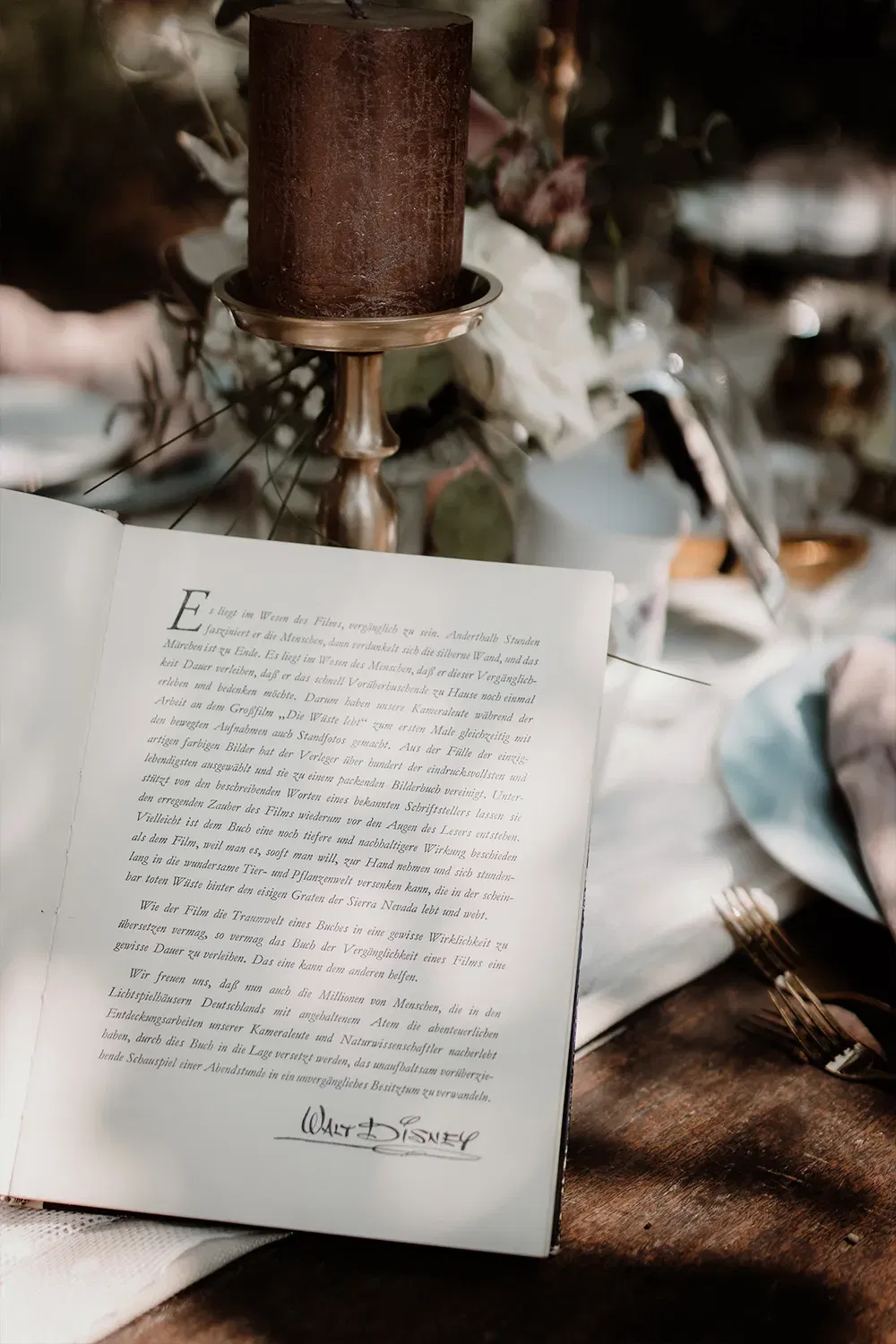 Ein aufgeschlagenes Buch mit einem Walt-Disney-Zitat liegt auf einem Holztisch, neben dekorativen Gegenständen wie einer Kerze und Blumen.
