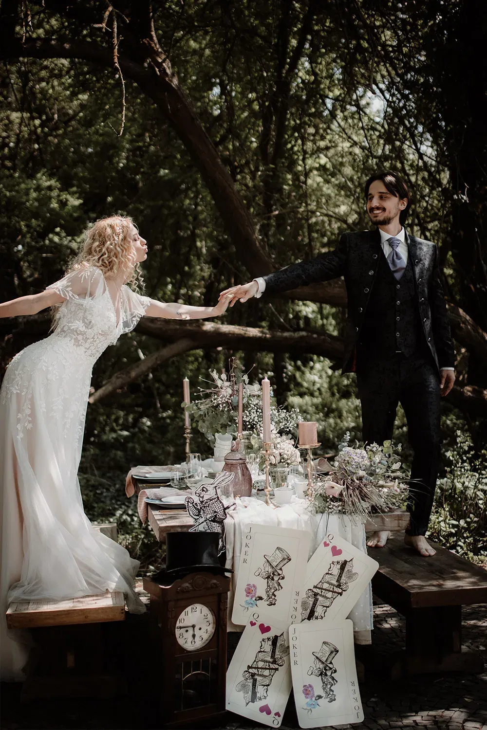 Die Braut und der Bräutigam strecken sich über einen festlich gedeckten Tisch die Hände entgegen. Der Tisch ist mit großen Spielkarten und antikem Dekor geschmückt.