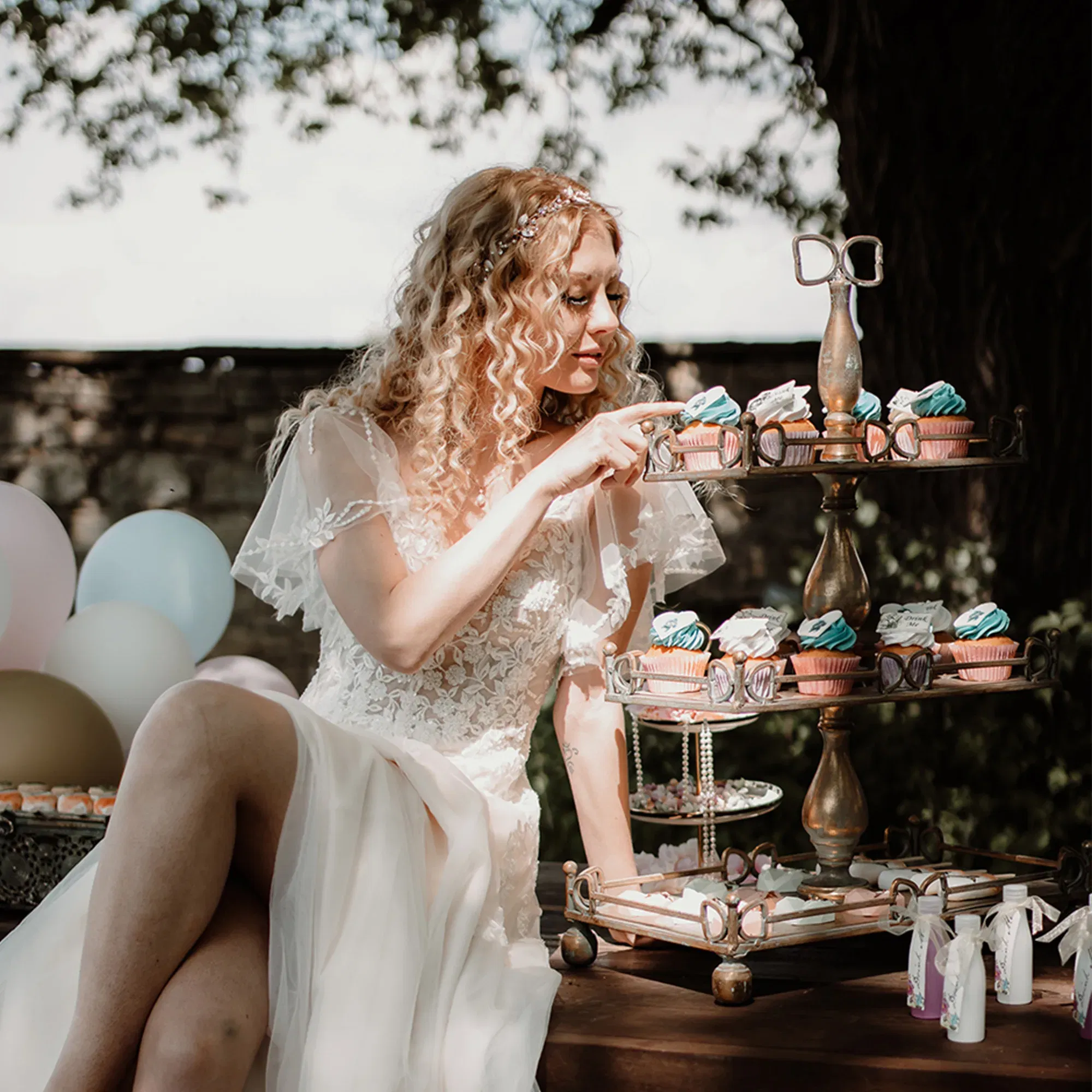 Eine Braut in einem eleganten weißen Kleid sitzt entspannt neben einer mehrstöckigen Etagere mit Cupcakes. Sie greift nach einer der süßen Köstlichkeiten, während hinter ihr bunte Luftballons den Rahmen bilden.