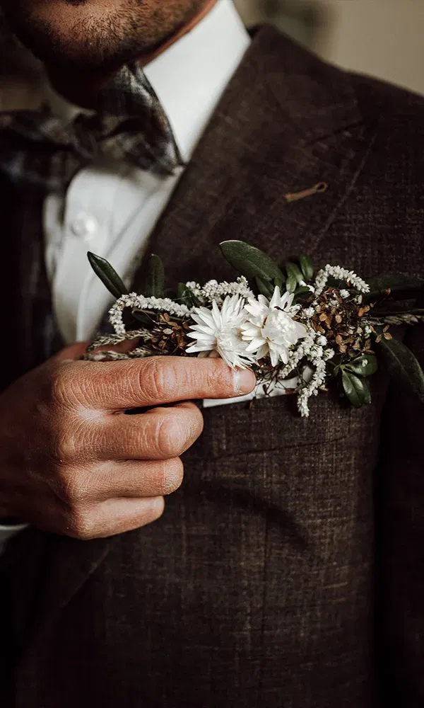 Nahaufnahme eines Bräutigams, der seinen filigranen Blumenanstecker mit weißen Blumen und dezentem Grün an seinem dunkelbraunen Anzug befestigt.