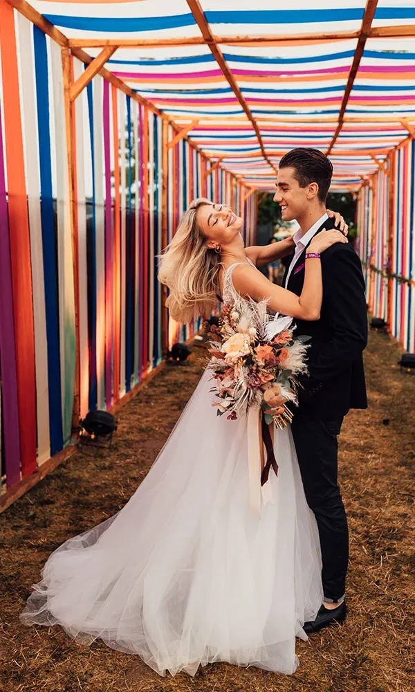 Ein frisch verheiratetes Paar posiert glücklich in einem Tunnel aus farbigen Stoffbahnen. Die Braut schwingt ihr Kleid, während sie von ihrem Bräutigam liebevoll gehalten wird. Beide wirken entspannt und voller Freude.