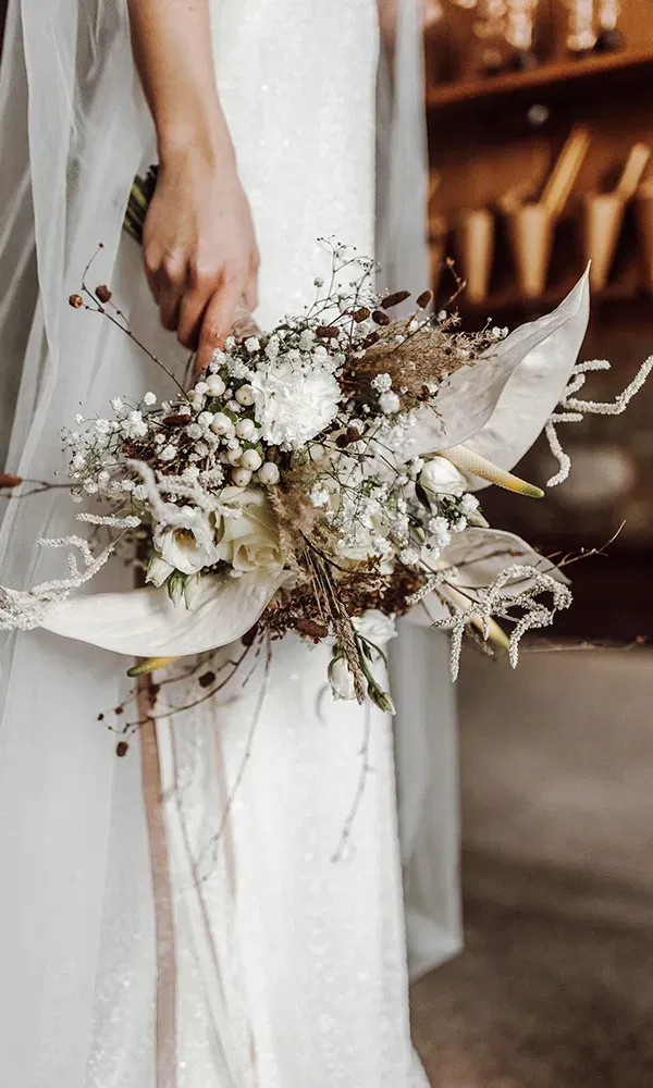 Nahaufnahme eines kunstvoll gestalteten Brautstraußes mit weißen Blumen und Naturmaterialien, der von der Braut in der Hand getragen wird.