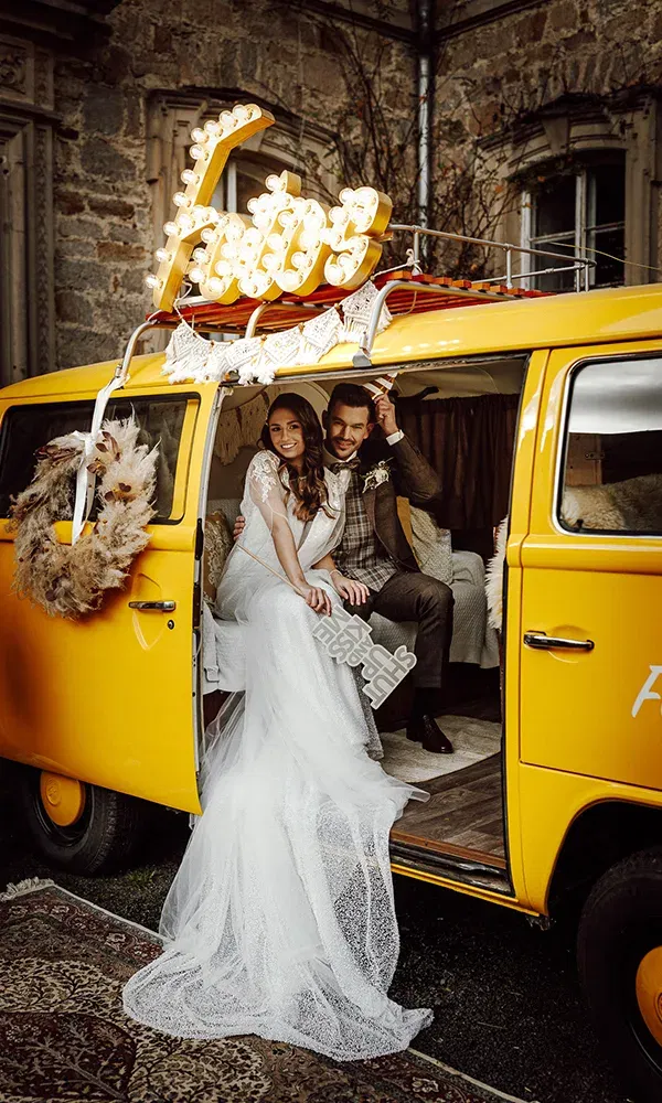 Ein fröhliches Hochzeitspaar posiert im Eingang eines gelben Vintage-Busses, verziert mit einem großen „Fotos“-Leuchtschild.
