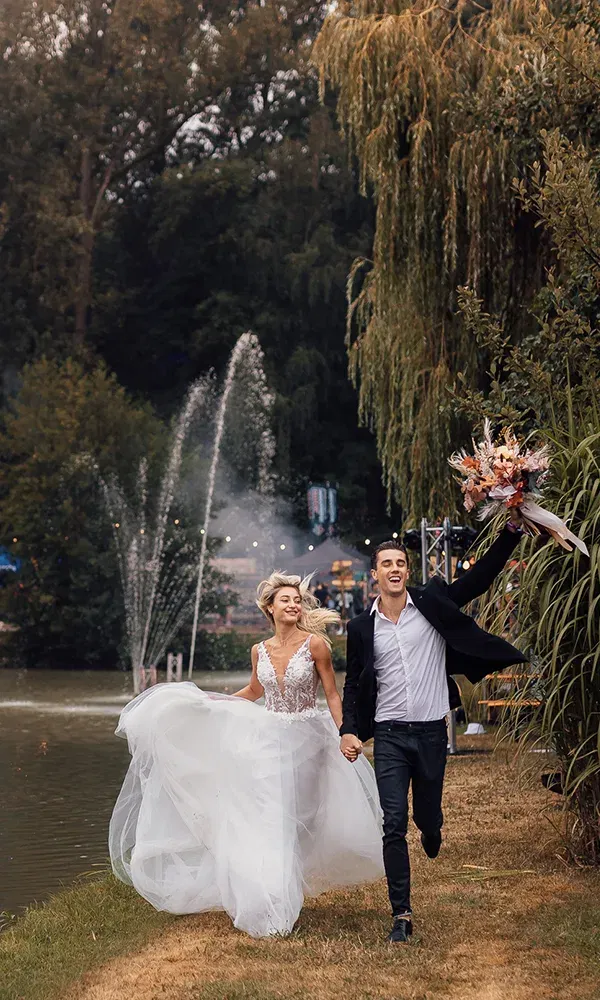 Das Brautpaar sprintet lachend entlang eines romantischen Sees mit Fontänen. Die Braut in ihrem fließenden Kleid und der Bräutigam in lässiger Kleidung strahlen Freude und Freiheit aus.