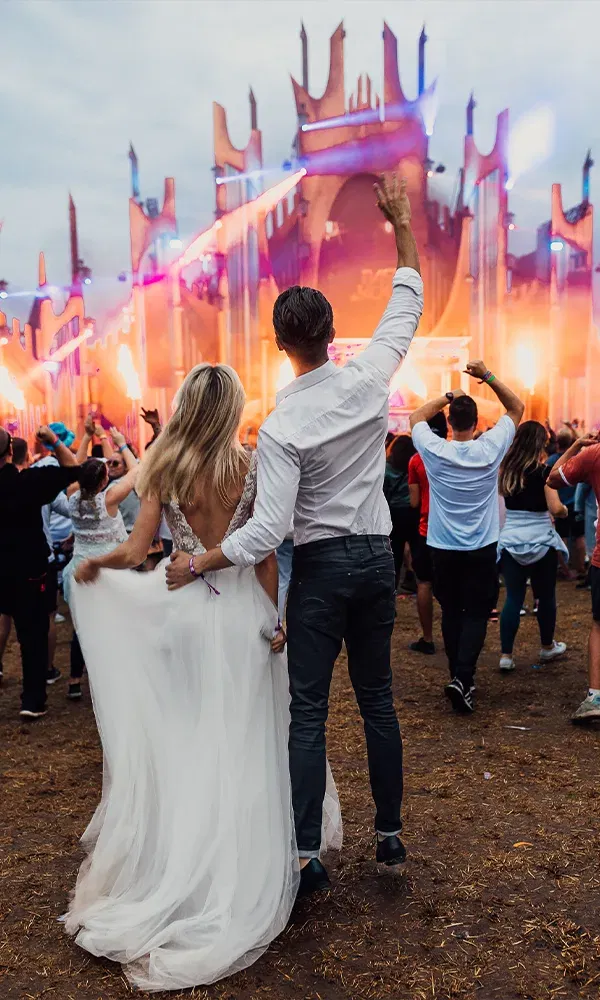 Ein junges Brautpaar steht Arm in Arm auf einem Festival und schaut auf die bunt beleuchtete Bühne. Die Braut hält ihren Schleier, und der Bräutigam hebt eine Hand hoch.