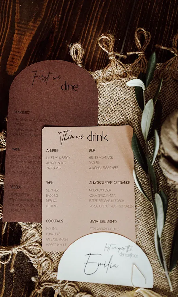 Menü- und Getränkekarten in stilvollem Design, platziert auf Jutestoff mit einer Olivenzweig-Dekoration, passend zur Hochzeitstafel.