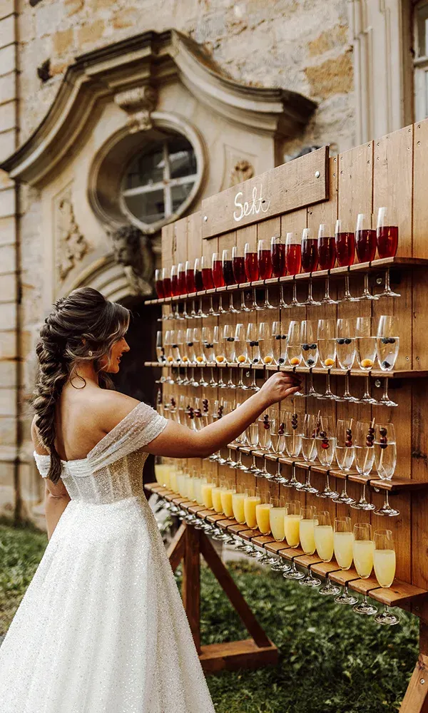 Die Braut, elegant gekleidet, nimmt ein Glas Sekt von einer dekorativen Sektwand während der Hochzeit.