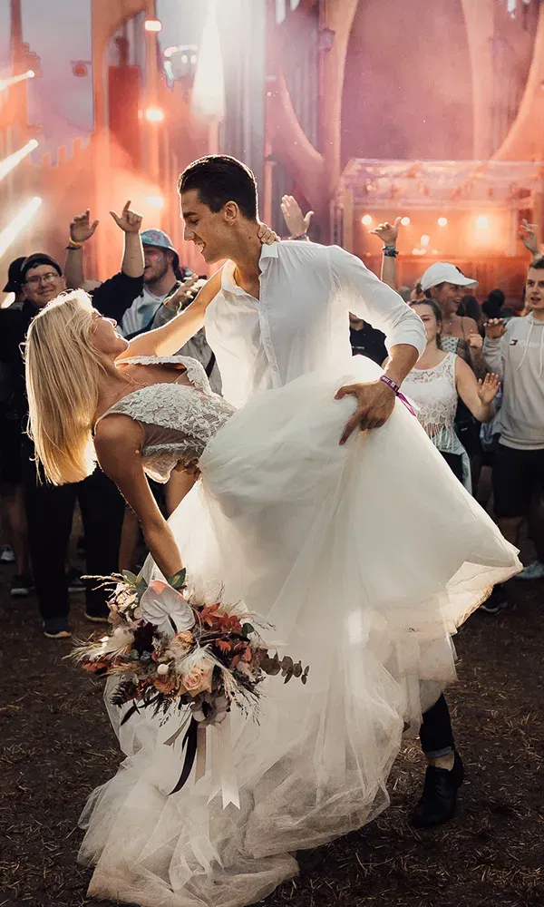 Ein Hochzeitspaar tanzt begeistert auf einem Festival, während der Bräutigam die Braut in seinen Armen hält. Der Hintergrund ist mit Feiernden und Lichtern beleuchtet.