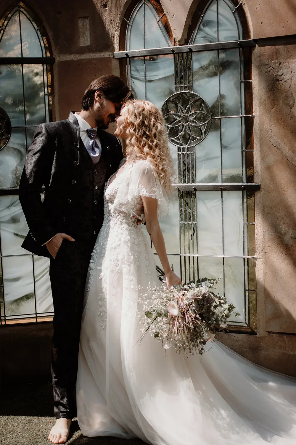 Eine Braut in einem eleganten Spitzenkleid steht dicht an ihrem Bräutigam, der einen schicken, schwarzen Anzug trägt. Das Paar lehnt sich aneinander, während sie vor einer kunstvoll gestalteten Glaswand posieren.