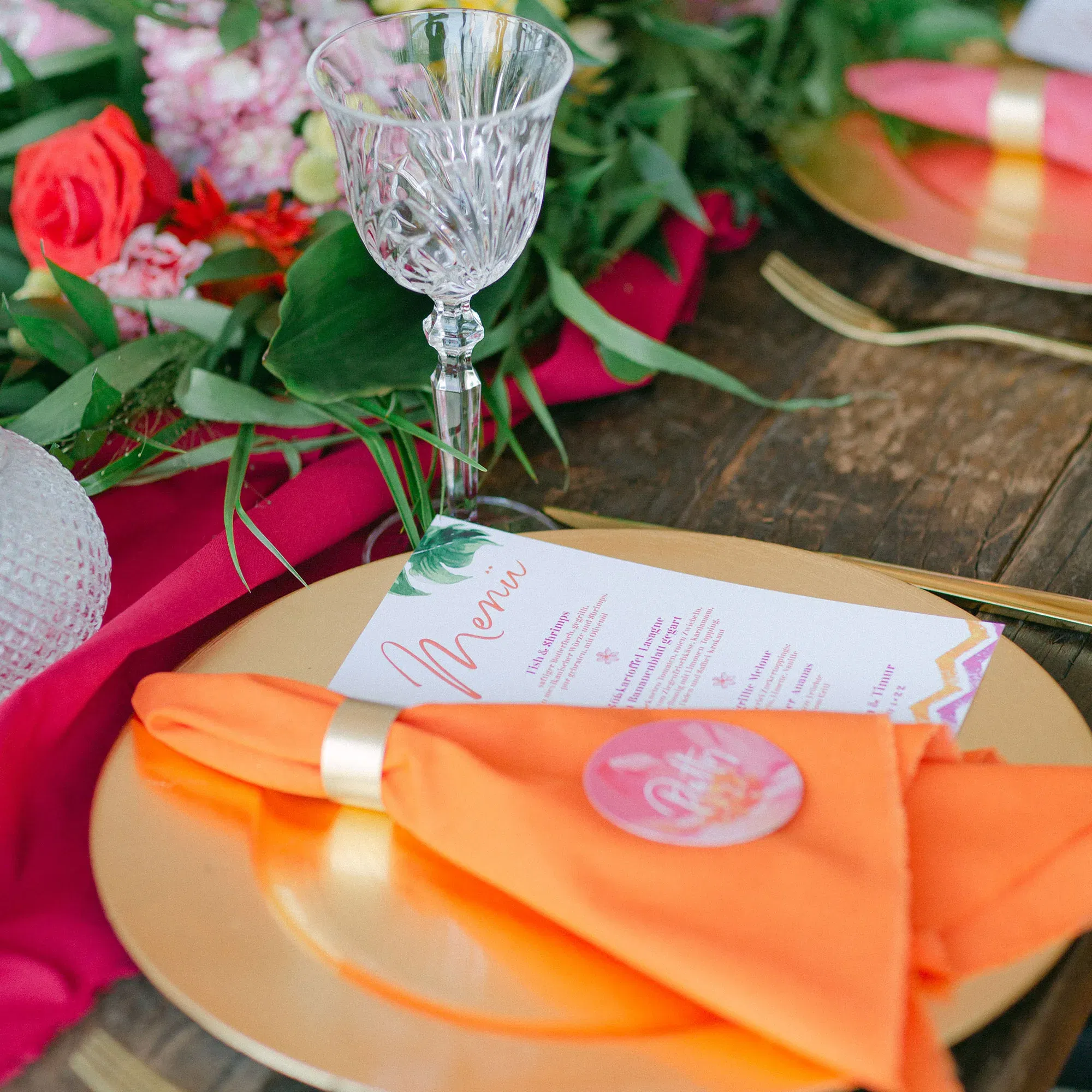 Eine bunte Hochzeitstafel, auf der eine orangefarbene Serviette mit einem goldenen Ring auf einem glänzenden goldenen Teller platziert ist. Ein filigranes Kristallglas und ein Menü in lebendigen Farben komplettieren das Bild.