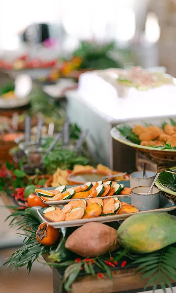 Eine reichhaltige Buffettauswahl mit tropischen Früchten wie Papaya und Süßkartoffeln, dazu gebackenes Gemüse auf einem Tablett, das von exotischen Blättern und frischen Zutaten umrahmt wird.