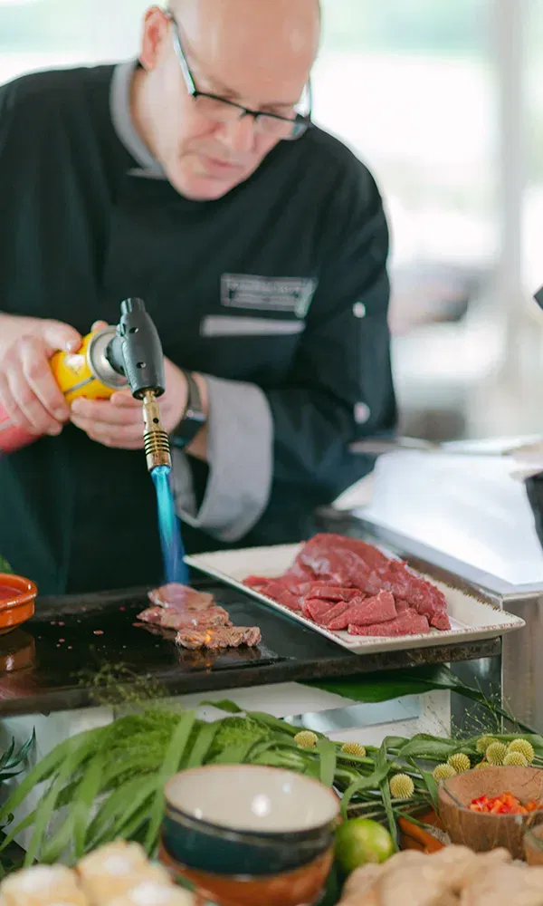 Nahaufnahme eines Kochs, der mit einem Flambierbrenner Fleisch auf einer Kochplatte zubereitet. Rundherum befinden sich verschiedene frische Zutaten wie Ingwer und Limetten.
