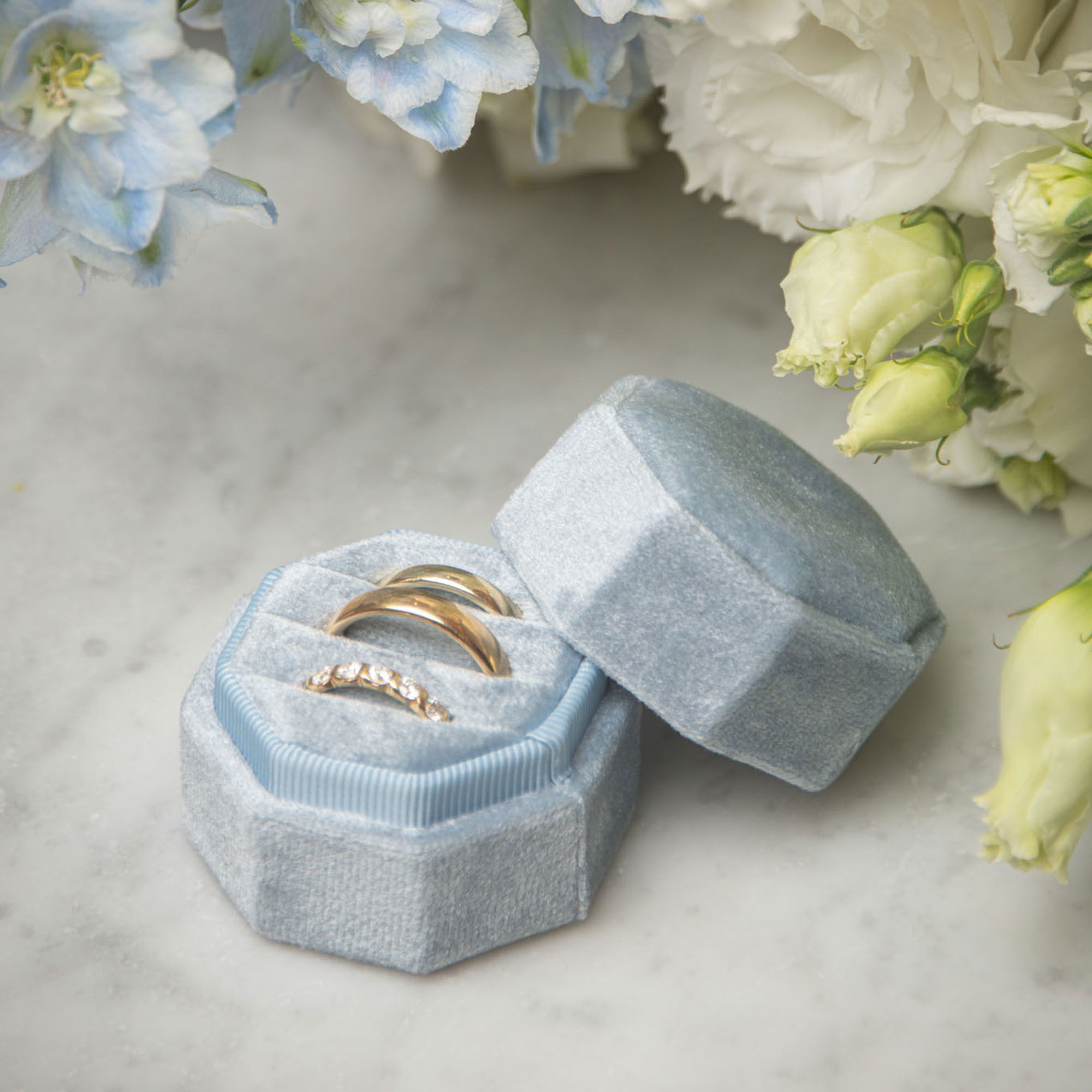 Ein luxuriöses blaues Schmuckkästchen enthält zwei goldene Eheringe, arrangiert neben weißen und blauen Blumen als Teil einer Hochzeitsdekoration.