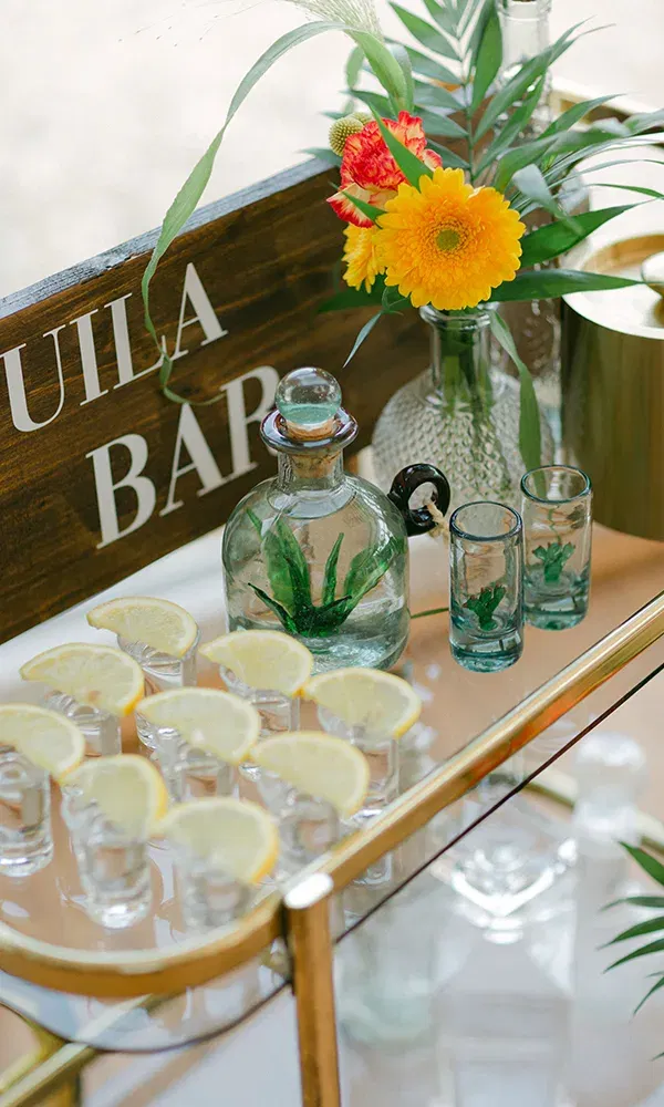 Elegantes Arrangement an einer Tequila-Bar, mit einer Tequilaflasche und dekorativen Shotgläsern, die mit Zitronenscheiben serviert werden. Umgeben von frischen Blumen und grünen Blättern.