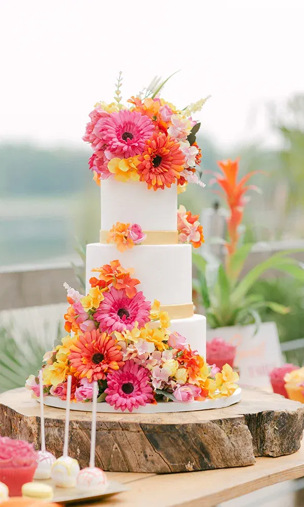 Eine elegante weiße Hochzeitstorte, geschmückt mit farbenfrohen Gänseblümchen in verschiedenen Pink- und Orangetönen. Die Blumen fließen von der obersten bis zur unteren Schicht der Torte.