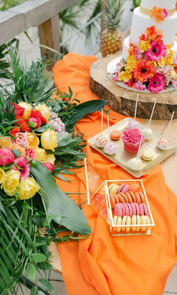 Auf einem Tisch sind pastellfarbene Macarons, Cupcakes und eine Hochzeitstorte mit farbenfrohen Blumen arrangiert, daneben liegen tropische Dekorationen aus frischen Blumen und Blättern.